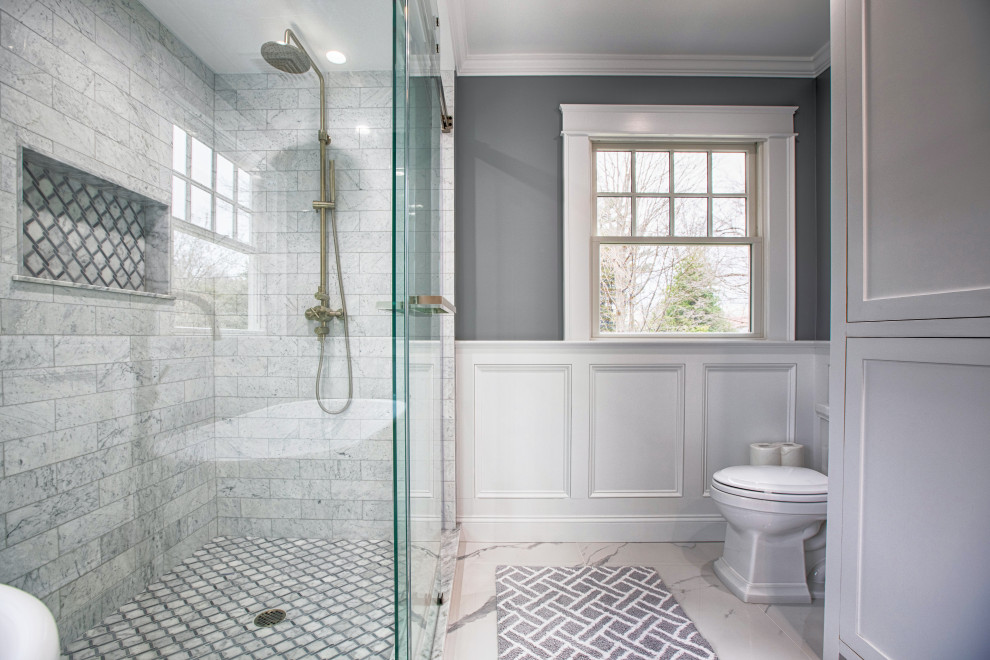 This is an example of a large transitional master bathroom in New York with shaker cabinets, white cabinets, a freestanding tub, an alcove shower, a one-piece toilet, white tile, marble, grey walls, porcelain floors, an undermount sink, marble benchtops, white floor, a sliding shower screen, white benchtops, a double vanity, a freestanding vanity and panelled walls.