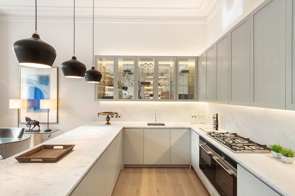 Photo of a contemporary u-shaped eat-in kitchen in London with an undermount sink, grey cabinets, white splashback, stainless steel appliances, light hardwood floors, a peninsula, brown floor and flat-panel cabinets.