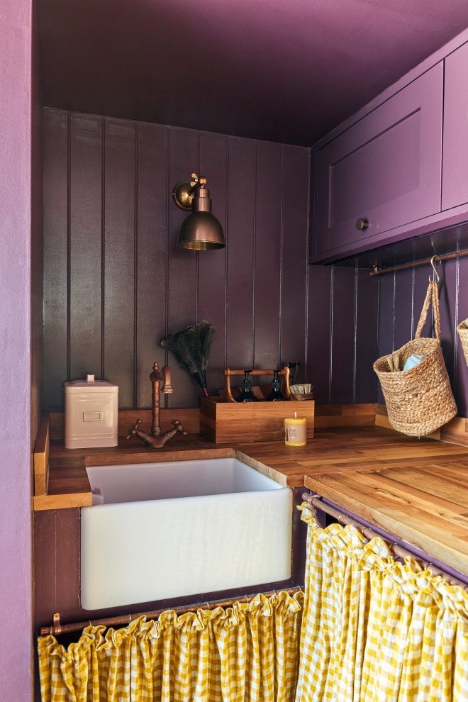 Photo of a mid-sized transitional l-shaped utility room in London with a farmhouse sink, shaker cabinets, yellow cabinets, wood benchtops, shiplap splashback, purple walls, limestone floors, a concealed washer and dryer, black floor, brown benchtop and planked wall panelling.