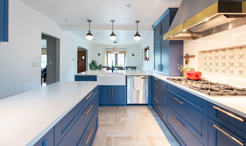 Réalisation d'une petite cuisine américaine tradition en U avec un évier de ferme, un placard à porte shaker, des portes de placard bleues, un plan de travail en quartz modifié, une crédence bleue, une crédence en céramique, un électroménager en acier inoxydable, un sol en carrelage de céramique, une péninsule, un sol marron et un plan de travail blanc.