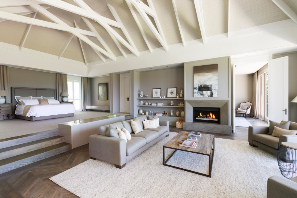 Expansive contemporary master bedroom in Melbourne with a stone fireplace surround, exposed beam, white walls, dark hardwood floors, a standard fireplace and brown floor.