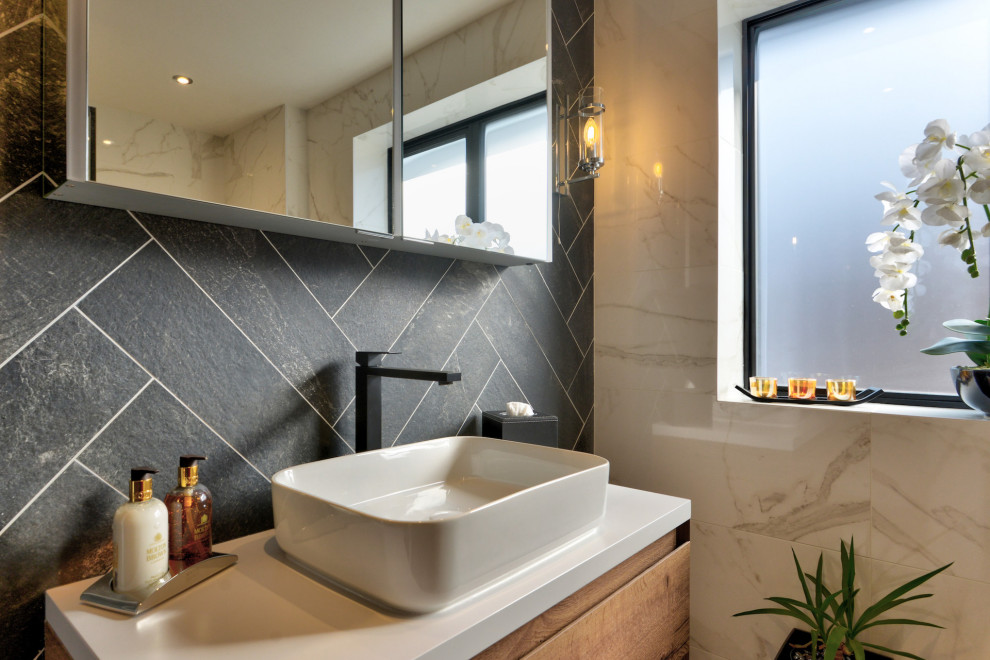 Photo of a medium sized contemporary grey and white family bathroom in Sussex with flat-panel cabinets, medium wood cabinets, a built-in bath, a shower/bath combination, a two-piece toilet, white tiles, marble tiles, grey walls, marble flooring, a console sink, quartz worktops, white floors, a hinged door, white worktops, a feature wall, a single sink and a floating vanity unit.