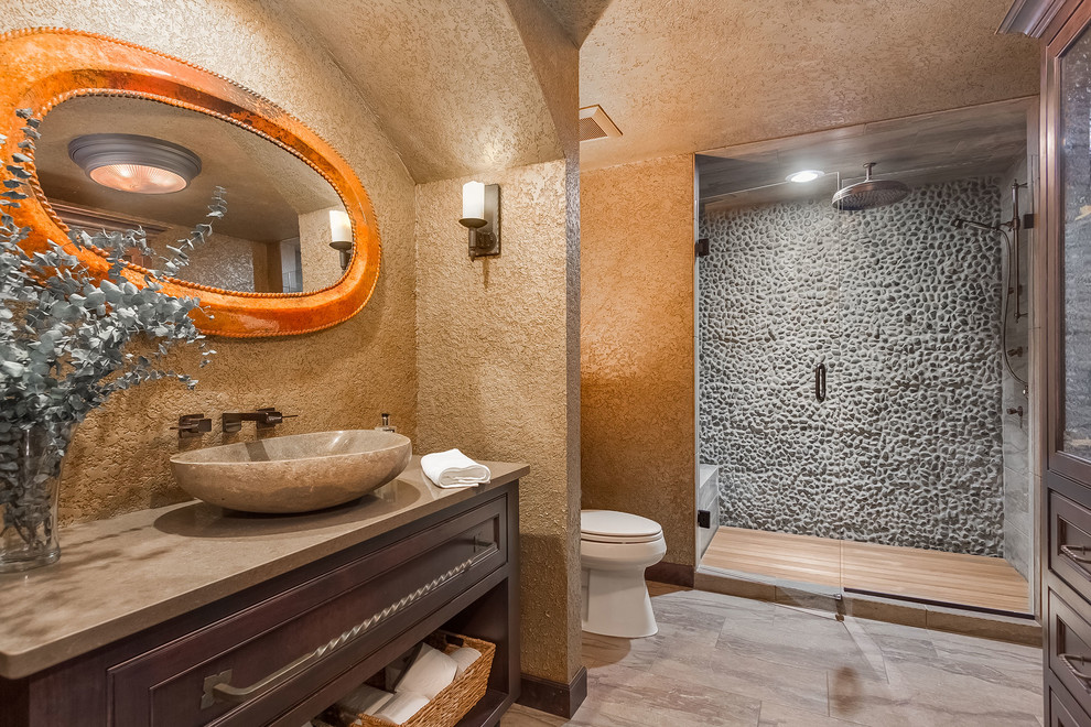Mid-sized traditional bathroom in Minneapolis with recessed-panel cabinets, dark wood cabinets, an alcove shower, a two-piece toilet, gray tile, pebble tile, brown walls, porcelain floors, a vessel sink, limestone benchtops, beige floor, a hinged shower door and brown benchtops.