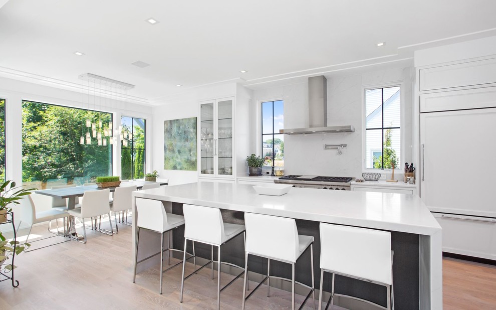 This is an example of a large beach style eat-in kitchen in New York with white cabinets, marble benchtops, white splashback, marble splashback, panelled appliances, light hardwood floors, with island, glass-front cabinets and beige floor.