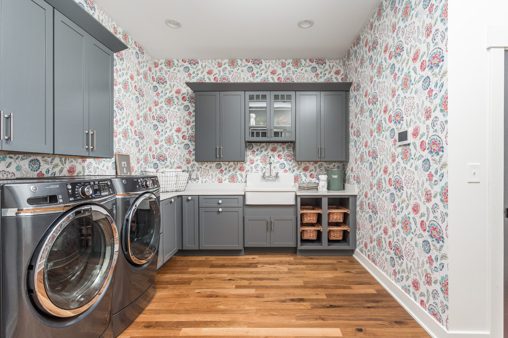 Inspiration for a large country l-shaped utility room in Indianapolis with a farmhouse sink, shaker cabinets, grey cabinets, multi-coloured walls, medium hardwood floors, a side-by-side washer and dryer, brown floor and white benchtop.
