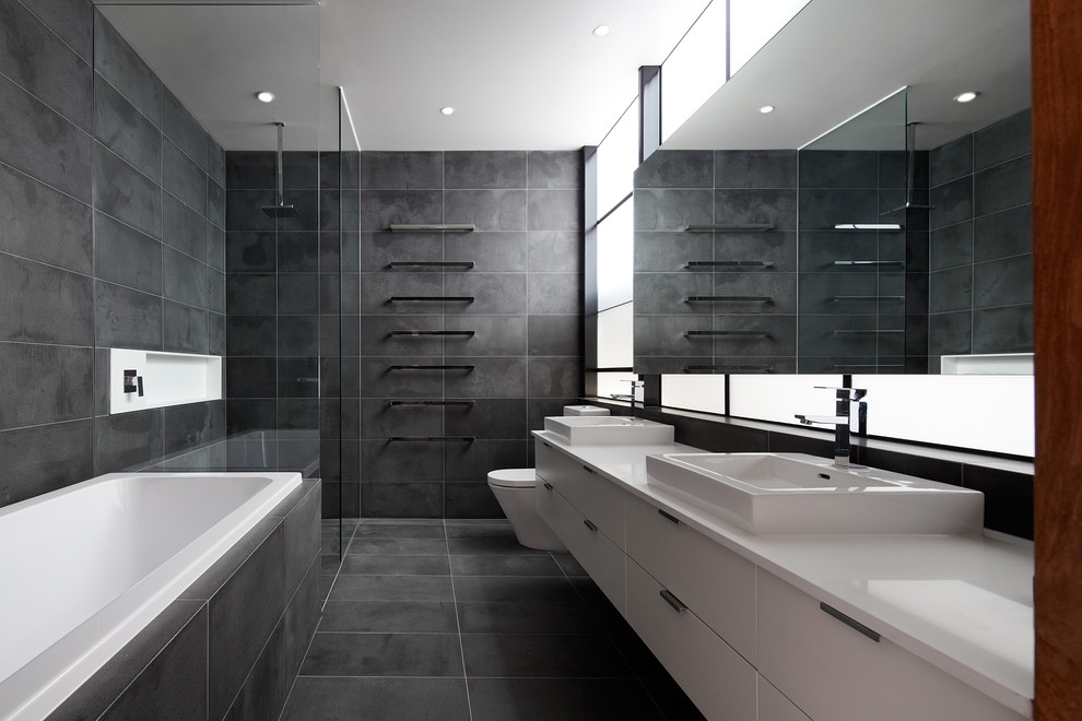 This is an example of an industrial bathroom in Melbourne with a console sink, flat-panel cabinets, white cabinets, engineered quartz benchtops, a drop-in tub, a curbless shower, a one-piece toilet, gray tile, stone tile and grey walls.