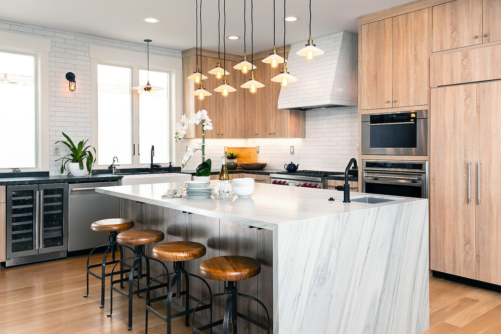 This is an example of a contemporary l-shaped kitchen in Other with a farmhouse sink, flat-panel cabinets, light wood cabinets, white splashback, stainless steel appliances, light hardwood floors, with island, brown floor and black benchtop.
