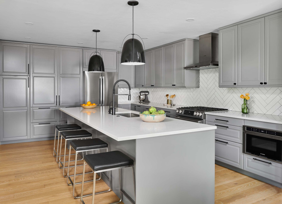 This is an example of a mid-sized transitional l-shaped eat-in kitchen in Denver with a single-bowl sink, flat-panel cabinets, grey cabinets, quartz benchtops, white splashback, ceramic splashback, stainless steel appliances, light hardwood floors, with island, brown floor and white benchtop.