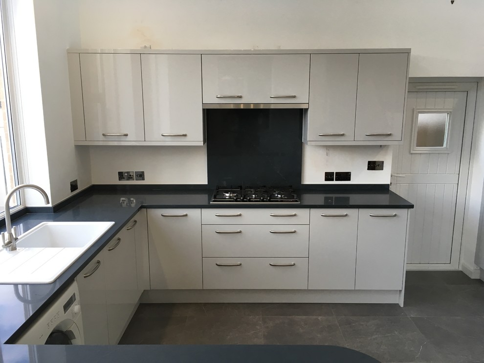 Photo of a modern u-shaped open plan kitchen in Oxfordshire with flat-panel cabinets, grey cabinets, solid surface benchtops, black splashback, white appliances, porcelain floors, grey floor and black benchtop.