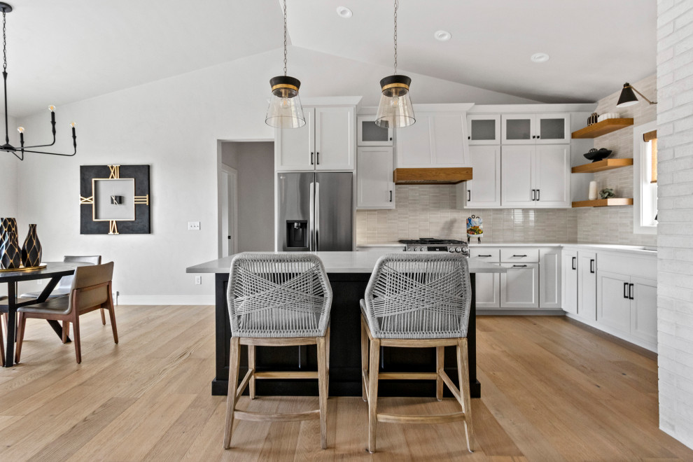 Example of a mid-sized transitional l-shaped medium tone wood floor, brown floor and vaulted ceiling eat-in kitchen design with an undermount sink, shaker cabinets, white cabinets, quartz countertops, white backsplash, porcelain backsplash, stainless steel appliances, an island and white countertops