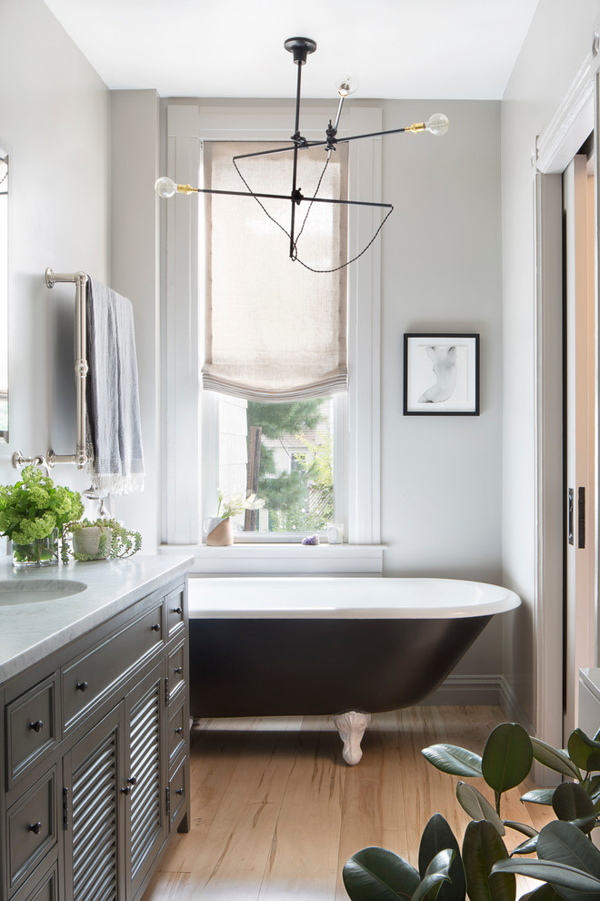Design ideas for a mid-sized eclectic master bathroom in New York with furniture-like cabinets, grey cabinets, a claw-foot tub, a two-piece toilet, white walls, medium hardwood floors, an undermount sink, marble benchtops and beige floor.