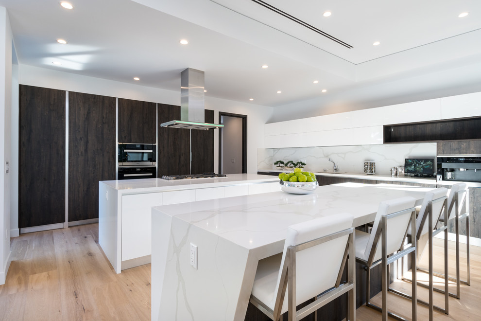 This is an example of a contemporary l-shaped separate kitchen in Los Angeles with flat-panel cabinets, white cabinets, quartz benchtops, white splashback, stone slab splashback, black appliances, light hardwood floors, multiple islands, beige floor and white benchtop.