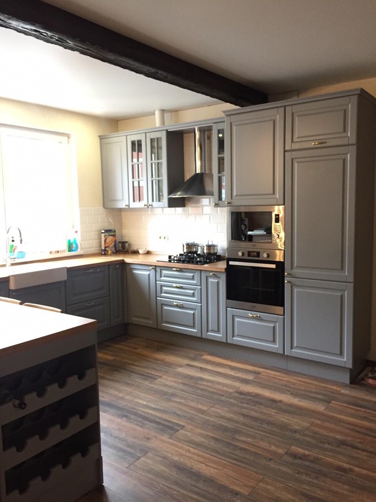 Photo of a large country u-shaped open plan kitchen in Other with a farmhouse sink, raised-panel cabinets, grey cabinets, laminate benchtops, white splashback, subway tile splashback, stainless steel appliances, with island and beige benchtop.