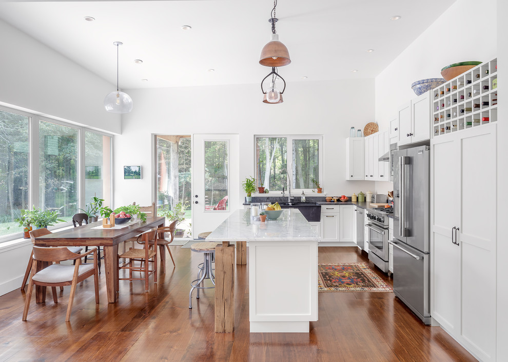This is an example of a transitional l-shaped eat-in kitchen in Portland Maine with white cabinets, stainless steel appliances, medium hardwood floors, with island, black benchtop, a farmhouse sink and shaker cabinets.