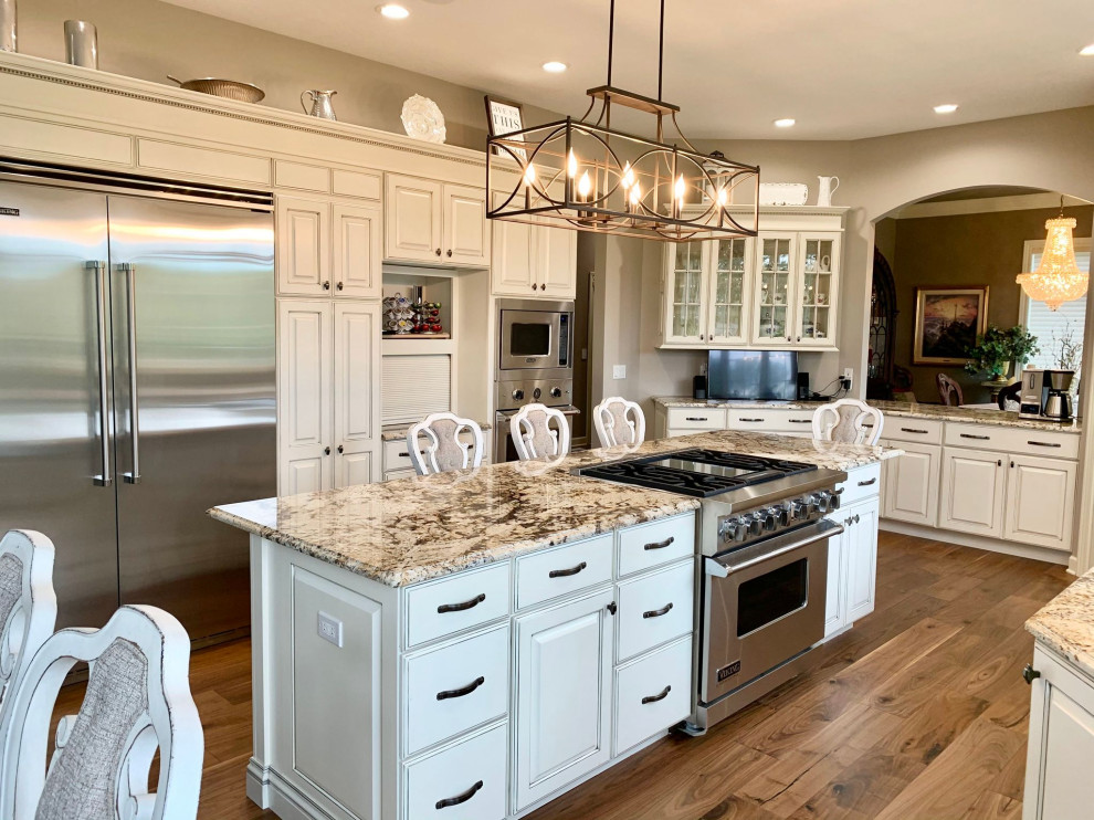 Large traditional u-shaped eat-in kitchen in Other with a farmhouse sink, raised-panel cabinets, white cabinets, granite benchtops, stainless steel appliances, medium hardwood floors, with island, brown floor and multi-coloured benchtop.