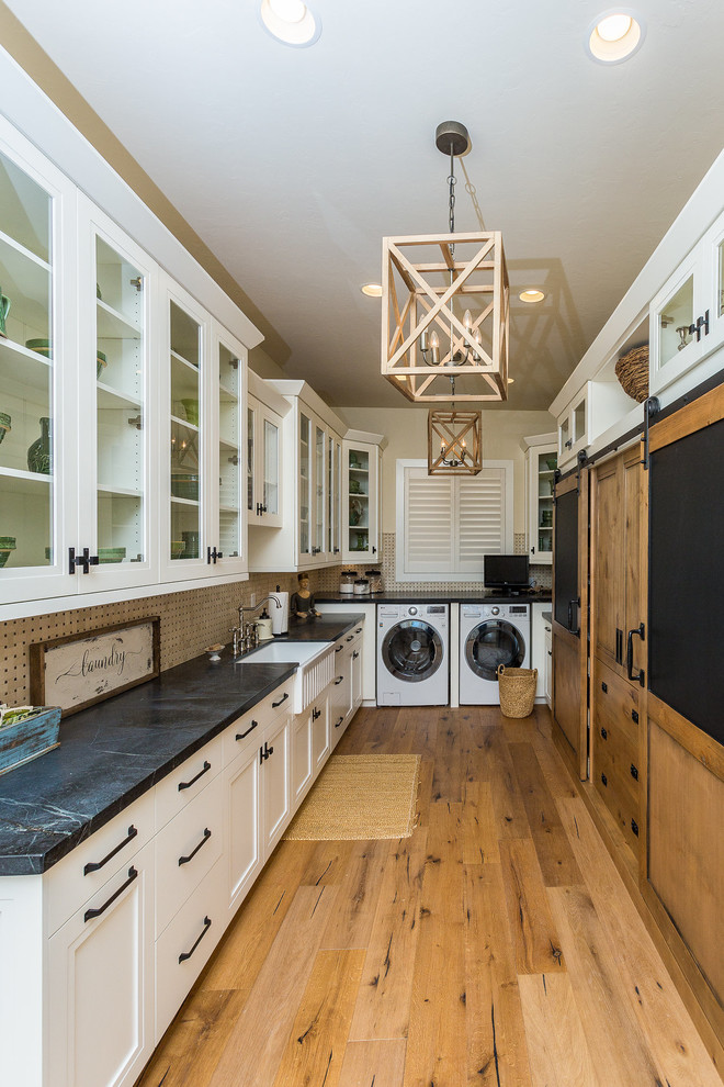 Large country u-shaped laundry room in Other with a farmhouse sink, shaker cabinets, white cabinets, soapstone benchtops, beige walls, medium hardwood floors, a side-by-side washer and dryer, brown floor and black benchtop.