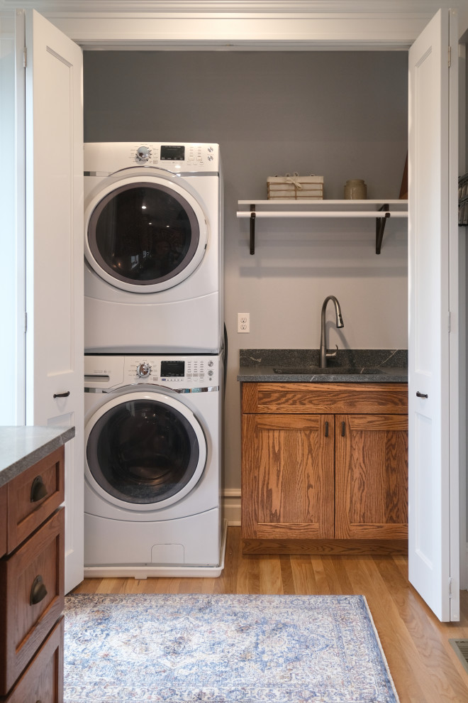 Photo of a mid-sized arts and crafts single-wall laundry cupboard in Baltimore with an undermount sink, shaker cabinets, brown cabinets, granite benchtops, grey walls, light hardwood floors, a stacked washer and dryer and grey benchtop.