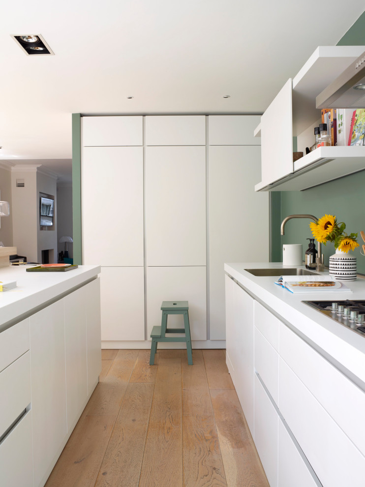 Large contemporary u-shaped open plan kitchen in London with an integrated sink, flat-panel cabinets, white cabinets, laminate benchtops, green splashback, stainless steel appliances, light hardwood floors, with island, beige floor and white benchtop.