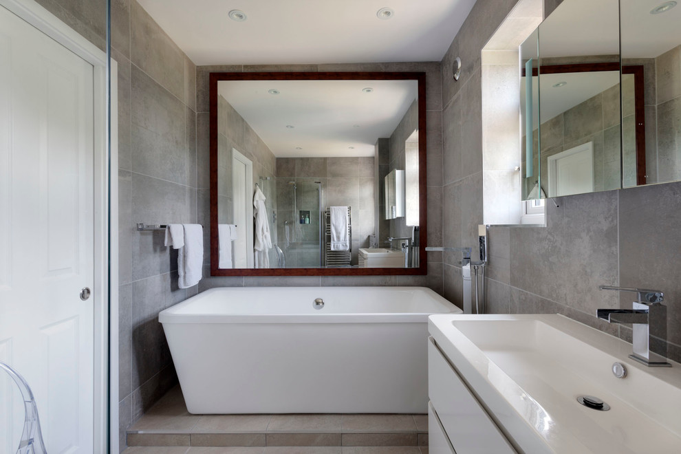 Contemporary bathroom in London with white cabinets, a freestanding tub, an open shower, ceramic tile, beige walls, a trough sink, flat-panel cabinets, solid surface benchtops, a wall-mount toilet, gray tile and ceramic floors.