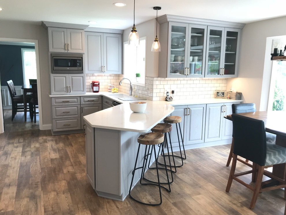 Mid-sized transitional u-shaped vinyl floor and brown floor enclosed kitchen photo with an undermount sink, raised-panel cabinets, gray cabinets, quartz countertops, white backsplash, ceramic backsplash, a peninsula and white countertops