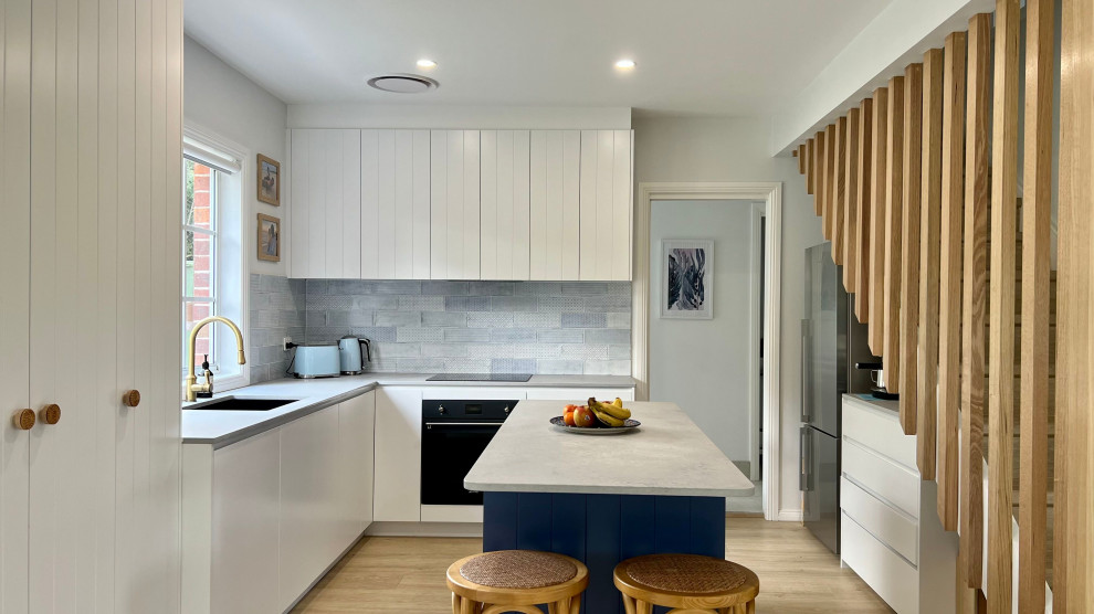 Design ideas for a mid-sized beach style l-shaped eat-in kitchen in Sydney with an undermount sink, recessed-panel cabinets, white cabinets, quartz benchtops, blue splashback, ceramic splashback, black appliances, light hardwood floors, with island, brown floor and grey benchtop.