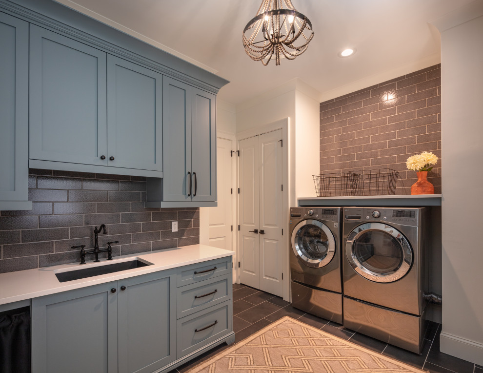 アトランタにある高級な広いトランジショナルスタイルのおしゃれな家事室 (アンダーカウンターシンク、シェーカースタイル扉のキャビネット、青いキャビネット、クオーツストーンカウンター、茶色いキッチンパネル、サブウェイタイルのキッチンパネル、白い壁、磁器タイルの床、左右配置の洗濯機・乾燥機、グレーの床、白いキッチンカウンター、白い天井) の写真