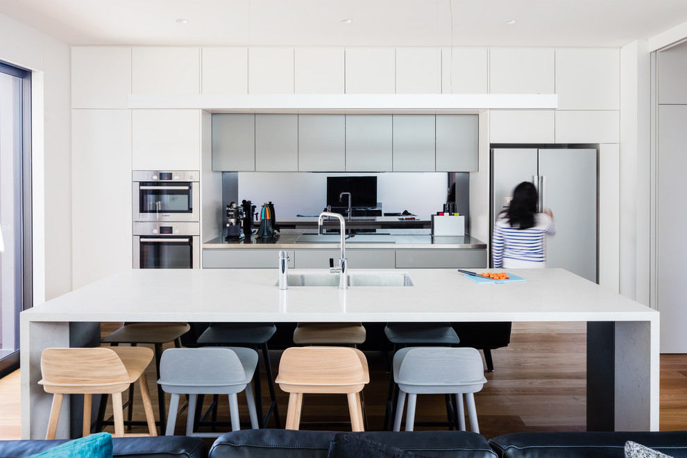 Inspiration for a contemporary galley kitchen in Sydney with white cabinets, with island, an undermount sink, flat-panel cabinets, mirror splashback, stainless steel appliances, medium hardwood floors, brown floor and white benchtop.