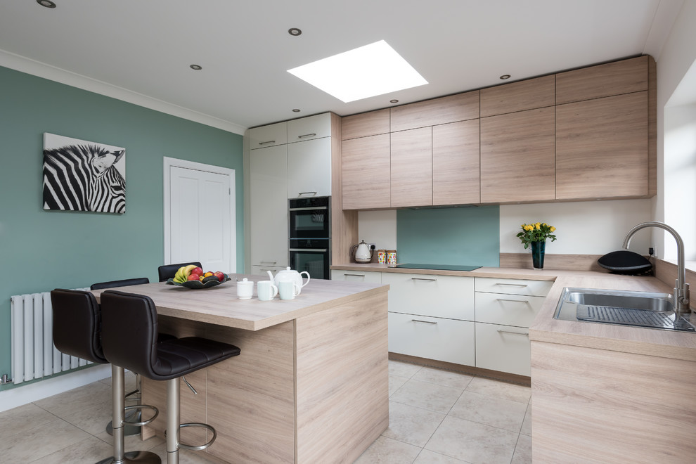 This is an example of a scandinavian l-shaped eat-in kitchen in Surrey with a single-bowl sink, flat-panel cabinets, light wood cabinets, laminate benchtops, green splashback, glass sheet splashback, black appliances, ceramic floors, with island, beige floor and beige benchtop.