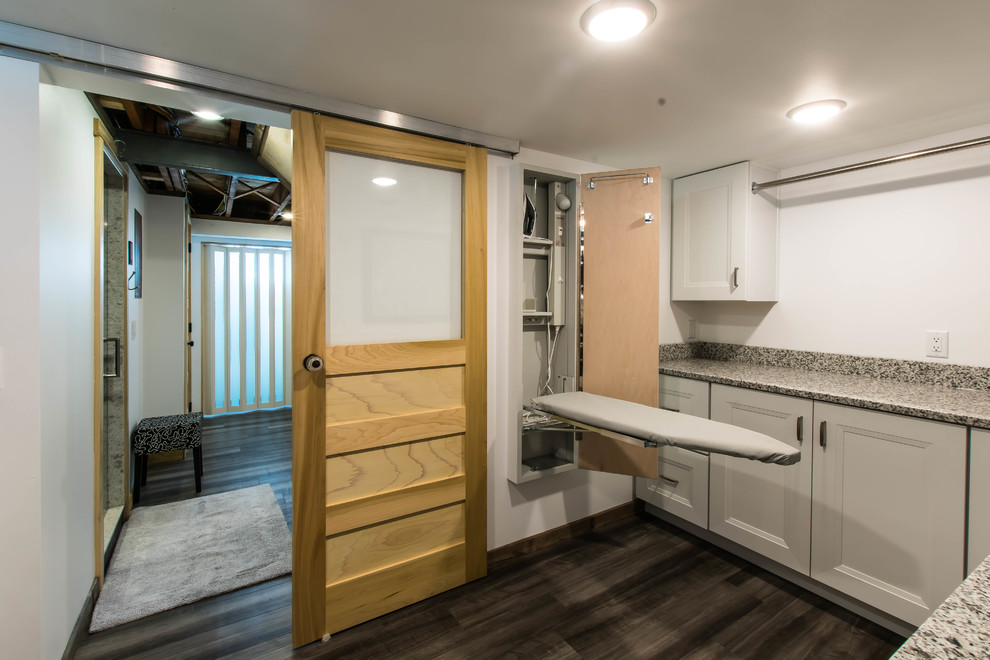Mid-sized country l-shaped dedicated laundry room in Other with an utility sink, shaker cabinets, white cabinets, granite benchtops, white walls, vinyl floors, a stacked washer and dryer, grey floor and multi-coloured benchtop.