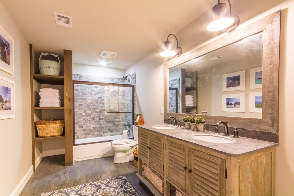 This is an example of a small country master bathroom in Atlanta with louvered cabinets, light wood cabinets, a drop-in tub, a shower/bathtub combo, a two-piece toilet, gray tile, stone tile, white walls, porcelain floors, an undermount sink, soapstone benchtops, grey floor, a sliding shower screen and grey benchtops.