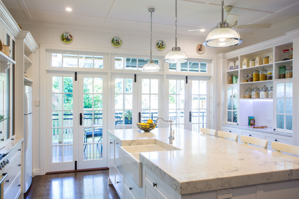 This is an example of a large traditional galley eat-in kitchen in Brisbane with a farmhouse sink, shaker cabinets, white cabinets, white splashback, subway tile splashback, with island, dark hardwood floors, white appliances and marble benchtops.