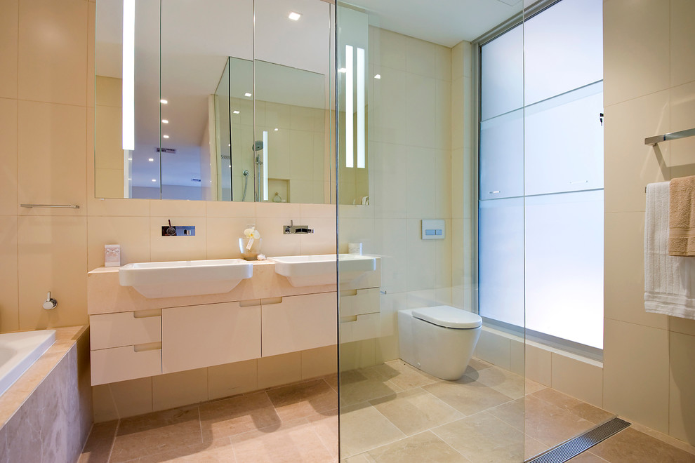 Contemporary bathroom in Other with limestone benchtops, limestone floors, a drop-in sink, beige cabinets, a drop-in tub, a curbless shower, a one-piece toilet, beige tile, stone tile, beige walls and flat-panel cabinets.