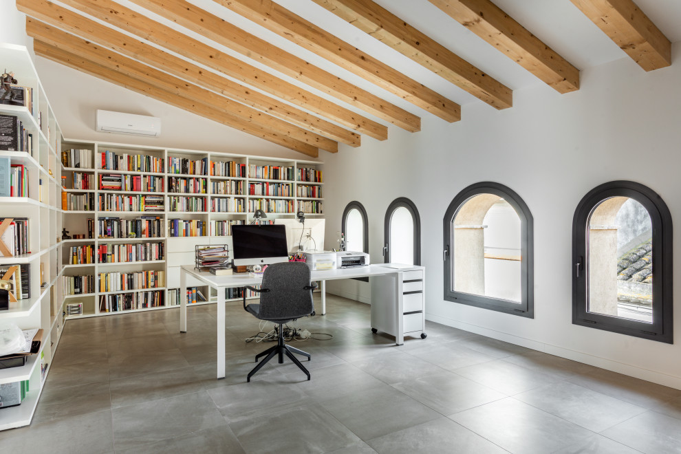 Modernes Arbeitszimmer mit weißer Wandfarbe, Porzellan-Bodenfliesen, freistehendem Schreibtisch, grauem Boden und freigelegten Dachbalken in Sonstige