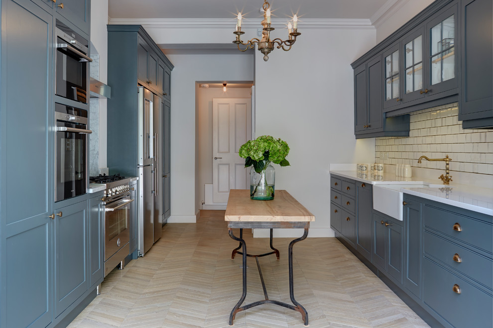 Design ideas for a large traditional galley open plan kitchen in London with a farmhouse sink, shaker cabinets, blue cabinets, quartzite benchtops, white splashback, ceramic splashback, stainless steel appliances, light hardwood floors, with island, brown floor and white benchtop.