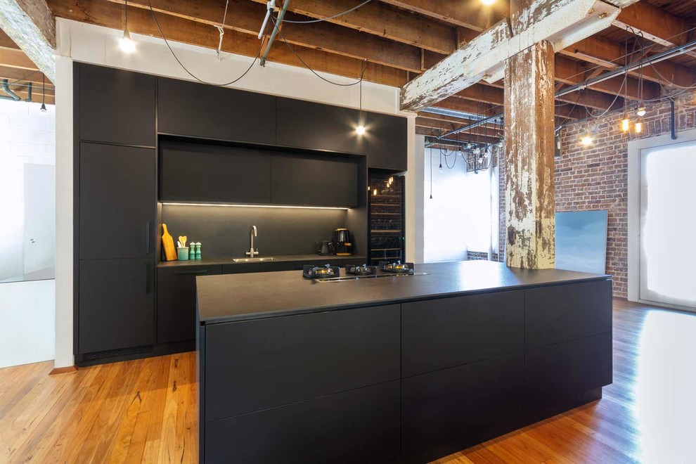 Small industrial galley eat-in kitchen in Sydney with an undermount sink, black cabinets, black splashback, panelled appliances, medium hardwood floors, with island, brown floor and black benchtop.