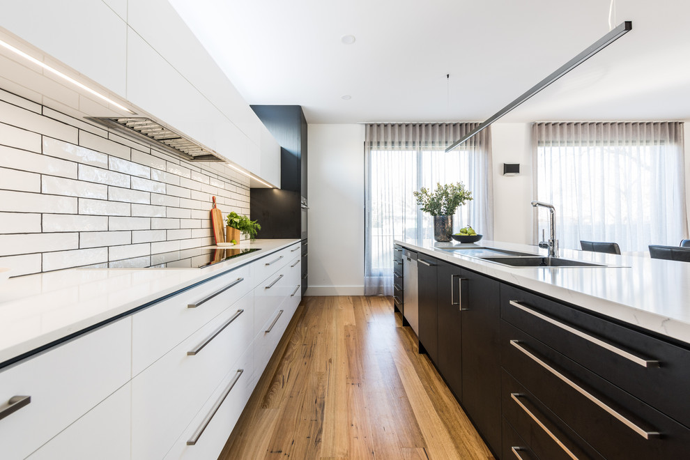 Inspiration for a large modern galley open plan kitchen in Melbourne with a double-bowl sink, flat-panel cabinets, black cabinets, quartz benchtops, white splashback, ceramic splashback, black appliances, light hardwood floors, with island, brown floor and white benchtop.