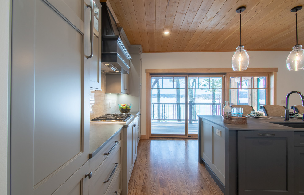 This is an example of a mid-sized country l-shaped eat-in kitchen in Boston with a single-bowl sink, shaker cabinets, grey cabinets, quartz benchtops, grey splashback, porcelain splashback, stainless steel appliances, medium hardwood floors, with island, brown floor and grey benchtop.