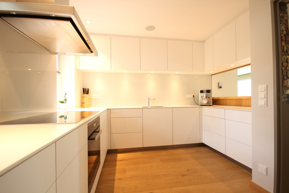 Photo of a mid-sized contemporary u-shaped separate kitchen in Munich with an undermount sink, flat-panel cabinets, white cabinets, solid surface benchtops, white splashback, glass sheet splashback, black appliances, medium hardwood floors, no island, brown floor and white benchtop.