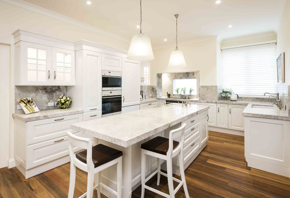 This is an example of a large traditional u-shaped eat-in kitchen in Melbourne with an undermount sink, raised-panel cabinets, white cabinets, marble benchtops, stone slab splashback, stainless steel appliances, medium hardwood floors, with island and grey splashback.