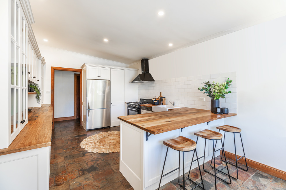 Photo of a mid-sized country l-shaped eat-in kitchen in Adelaide with a farmhouse sink, shaker cabinets, white cabinets, wood benchtops, white splashback, subway tile splashback, black appliances, slate floors, a peninsula, grey floor and yellow benchtop.