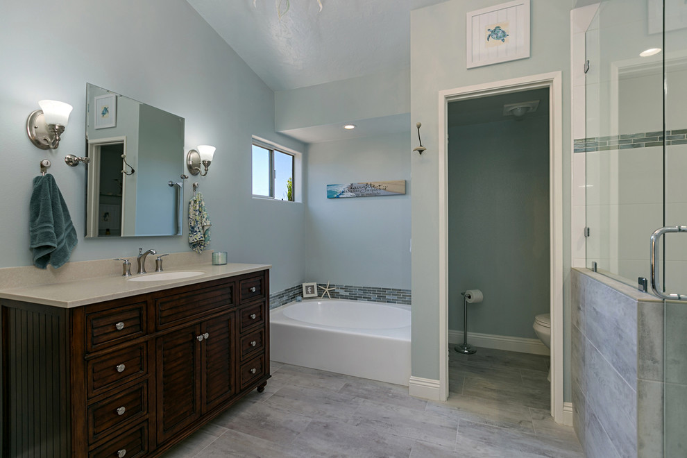 Photo of a large beach style master bathroom in San Diego with furniture-like cabinets, medium wood cabinets, a corner tub, a corner shower, a two-piece toilet, gray tile, porcelain tile, grey walls, porcelain floors, a drop-in sink, granite benchtops, grey floor and a hinged shower door.