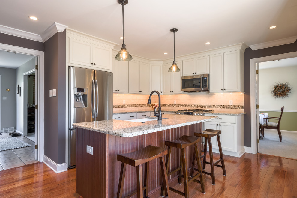 Photo of a mid-sized transitional u-shaped eat-in kitchen in Philadelphia with a farmhouse sink, recessed-panel cabinets, white cabinets, granite benchtops, subway tile splashback, stainless steel appliances, medium hardwood floors, with island, brown floor, white splashback and multi-coloured benchtop.