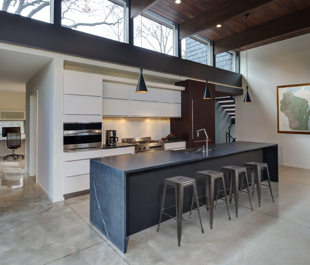 This is an example of a contemporary open plan kitchen in Milwaukee with a farmhouse sink, flat-panel cabinets, white cabinets, soapstone benchtops, stainless steel appliances, concrete floors, with island and grey floor.