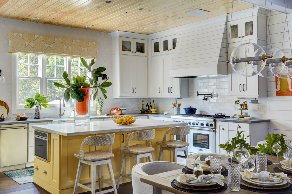 This is an example of a country l-shaped eat-in kitchen in Houston with a farmhouse sink, recessed-panel cabinets, yellow cabinets, white splashback, subway tile splashback, stainless steel appliances, medium hardwood floors, with island, brown floor, quartz benchtops and white benchtop.