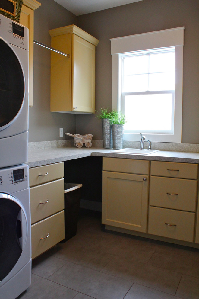 Photo of a laundry room in Grand Rapids with a drop-in sink, flat-panel cabinets, yellow cabinets, grey walls, porcelain floors and a stacked washer and dryer.