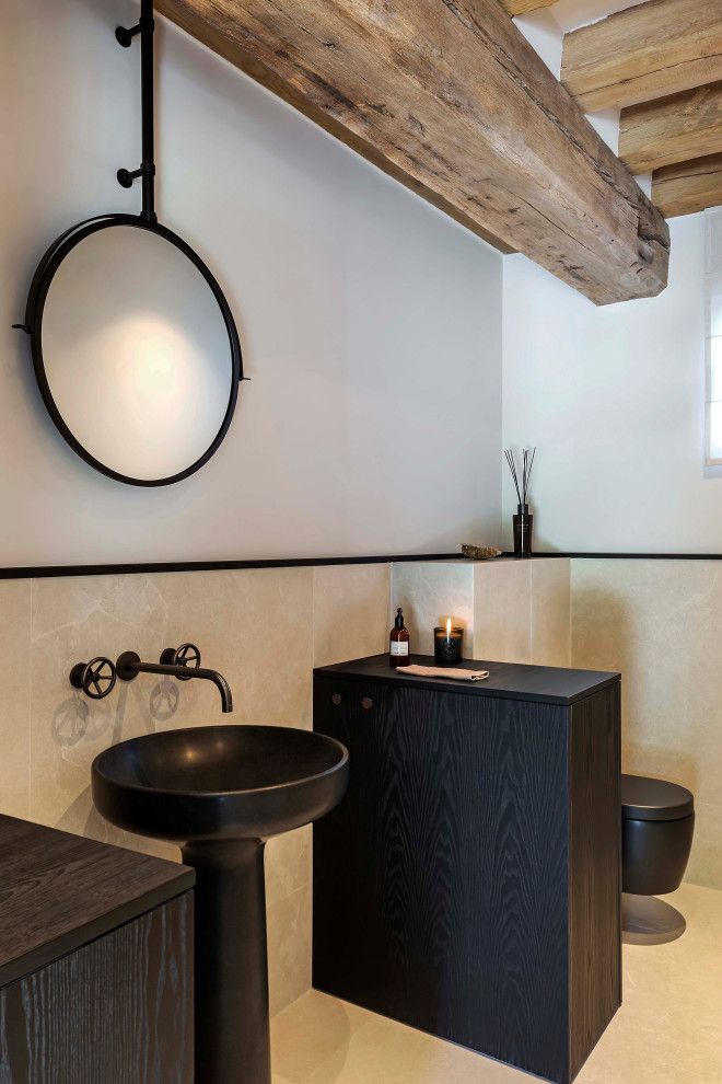 Bild på ett stort industriellt toalett, med svarta skåp, en vägghängd toalettstol, beige kakel, kakelplattor, beige väggar, kalkstensgolv, ett nedsänkt handfat, marmorbänkskiva och beiget golv