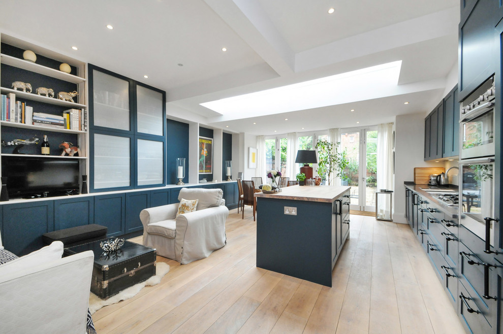 This is an example of a large eclectic eat-in kitchen in London with a double-bowl sink, raised-panel cabinets, blue cabinets, wood benchtops, stainless steel appliances, light hardwood floors and with island.
