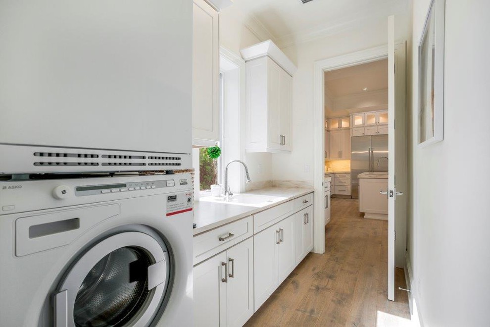 Large beach style galley utility room in Miami with a drop-in sink, shaker cabinets, white cabinets, quartz benchtops, white walls, medium hardwood floors and a stacked washer and dryer.