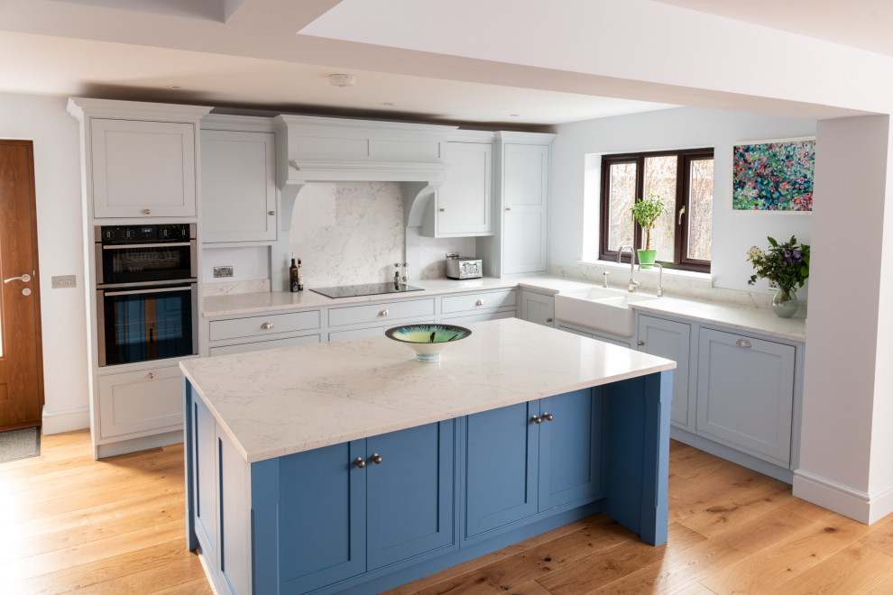 Large beach style l-shaped open plan kitchen in Essex with a farmhouse sink, shaker cabinets, blue cabinets, quartzite benchtops, white splashback, stainless steel appliances, medium hardwood floors, with island, brown floor and white benchtop.