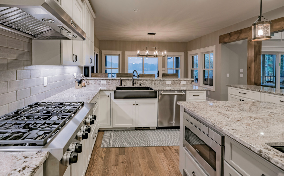 Large country l-shaped open plan kitchen in Atlanta with a farmhouse sink, shaker cabinets, white cabinets, granite benchtops, grey splashback, ceramic splashback, stainless steel appliances, medium hardwood floors, multiple islands, brown floor and multi-coloured benchtop.